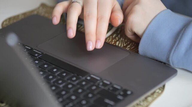 Close-up of woman's hands using laptop touchpad. Fingers glide smoothly across dark gray surface. Light blue sleeve visible. Hand wears simple ring. Background shows textured woven mat
