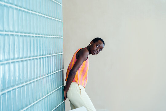 Curious Black woman tilting head near retro glass block wall, direct eye contact with questioning emotion, creative portrait, individuality, modern interior, urban lifestyle, minimal aesthetic