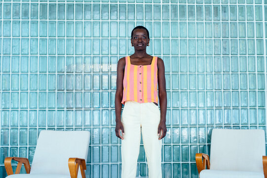 Confident Black woman standing between chairs against retro glass block wall, direct posture with calm strong emotion, modern interior design concept, symmetry composition, urban lifestyle