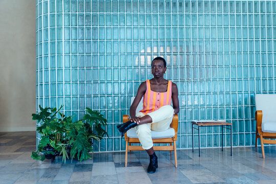 Calm Black woman sitting on chair with crossed legs, relaxed confident posture, minimalist retro interior with glass block wall, quiet moment, urban lifestyle, modern design space