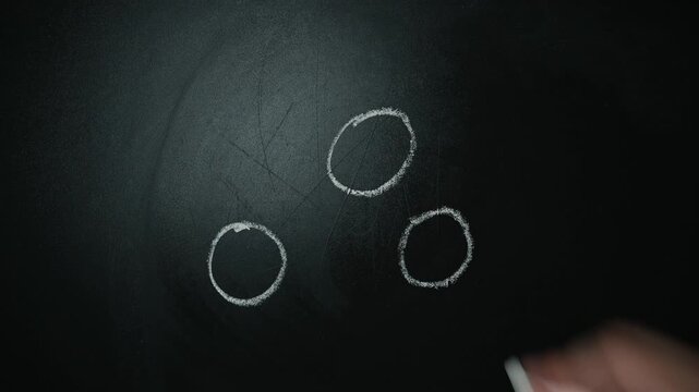 A person is using white chalk to draw several small circles on a dark chalkboard.