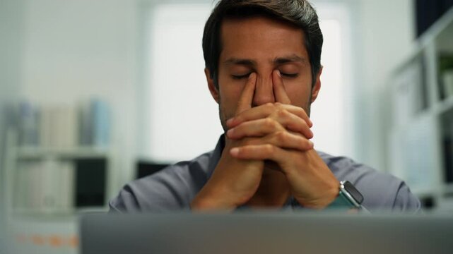 Hispanic man indoors, multitasking between computer and documents, exhausted from job pressure.