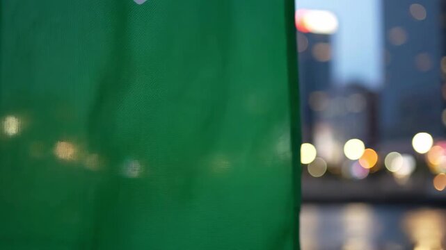 A green flag waving in front of a blurred cityscape at dusk, with a bokeh effect creating a sense of depth and atmosphere.