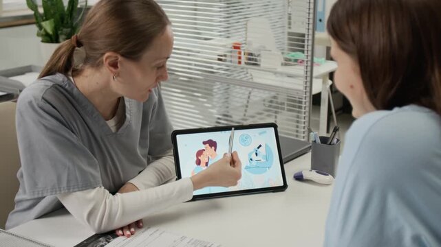 Zoom out view of fertility specialist pointing at digital tablet displaying IVF illustration while explaining procedure to young woman sitting in front of her