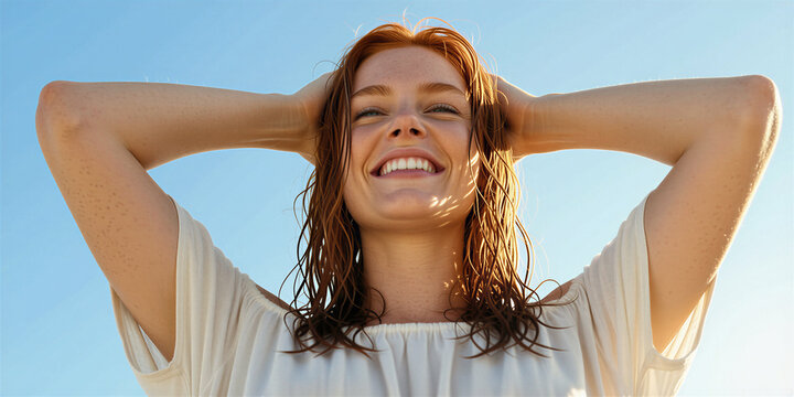 Slender red-haired woman in white shirt standing against blue sky, hands in hair, squinting and smiling, with sunny atmosphere.