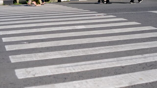 Pedestrian walks across zebra crossing. Sunlight casts long shadow on pavement. Casual outfit, white sneakers, brown pants. No emotion visible, focused on crossing. Ideal for urban, daily life