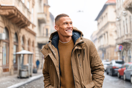 A smiling man in a brown jacket walks down a stylish city street on a cold day, surrounded by historic buildings and soft snowfall.