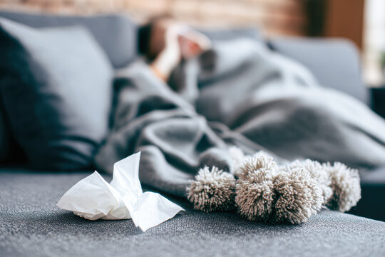 A person lies under a blanket on a couch with used tissue and a winter hat in the foreground, suggesting illness or rest.