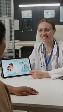 Vertical over the shoulder view of female fertility expert in lab coat pointing at illustration on digital tablet while providing assistance to young woman interested in IVF treatment