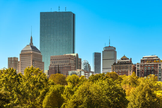High rise office towers with reflective glass and historic stone structures line the edge of the Boston skyline seen from a Boston Common park.