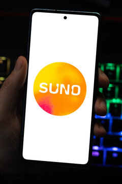 Punta Arenas, Chile &ndash; April 30, 2026: Close-up of the Suno artificial intelligence music generation brand logo on a mobile phone screen held by a hand over a dark desk with glowing RGB keyboard lights