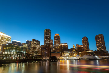 Fototapeta premium High-rise buildings glow against the dark blue evening sky over the canal in downtown Boston as seen from the adjacent Seaport district.