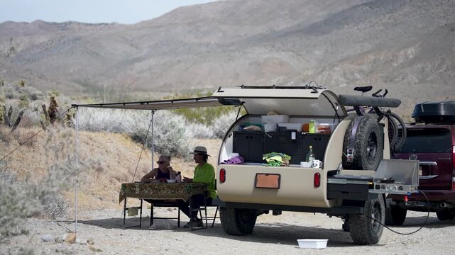 Mature couple enjoys a meal beside their teardrop camper trailer under an awning in the desert landscape of Plum Canyon.