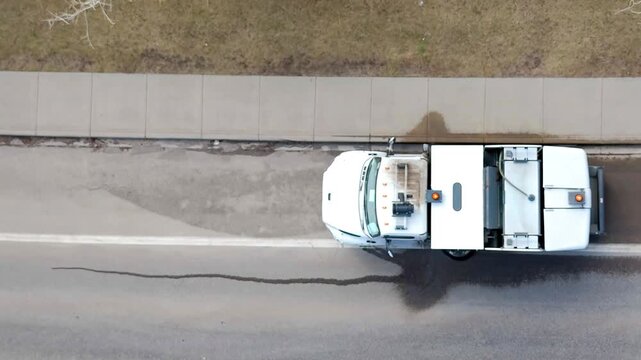 Aerial top-down view of a white street sweeper truck cleaning a suburban asphalt road next to a concrete sidewalk during the day.