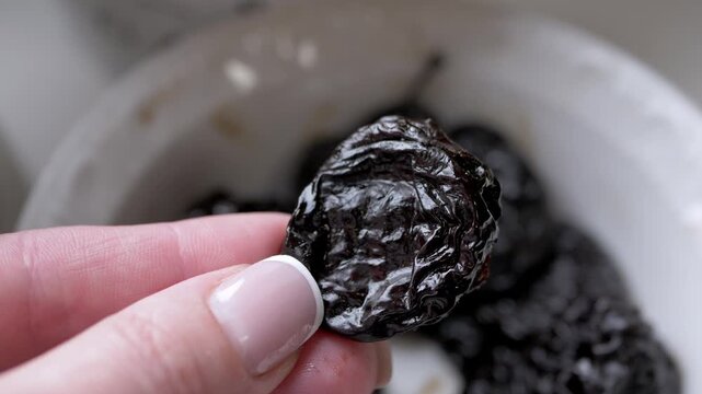 A woman hand holds a single glossy dried prune over a ceramic bowl filled with more prunes in a kitchen. Concept of healthy snacking, natural fiber, and organic dried fruit nutrition.