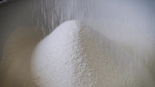 White wheat flour falls steadily into a growing pile from an unseen sifter, creating a soft, powdery texture in a kitchen setting. Concept of culinary arts, home baking, and precision.