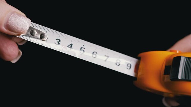Female hands with manicured nails pull out a yellow measuring tape over a black background. Concept of precision measurement, tailoring accuracy, and home improvement DIY projects.