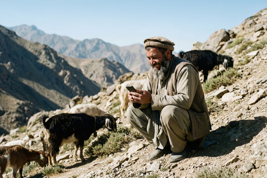 A laughing shepherd using a smartphone among a herd of goats in the mountains