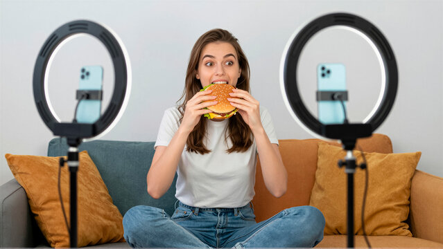 Young woman influencer holds a large burger while filming a video for social media. She sits on a sofa between two ring light setups with a smartphone in a bright living room