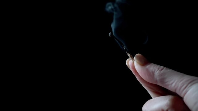 A person holds a recently extinguished matchstick with grey smoke billowing upwards against a solid black background. Side view. Concept of psychological burnout, exhaustion, and the fading of energy.