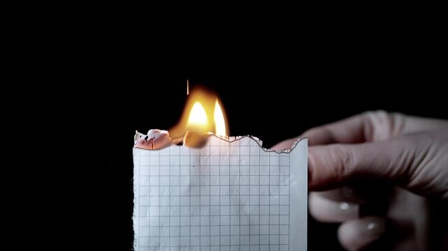 A hand holds a piece of grid paper and ignites the edge with a lit match in complete darkness. Concept of destroying evidence, burning secret documents, forgetting ideas, and erasing past.