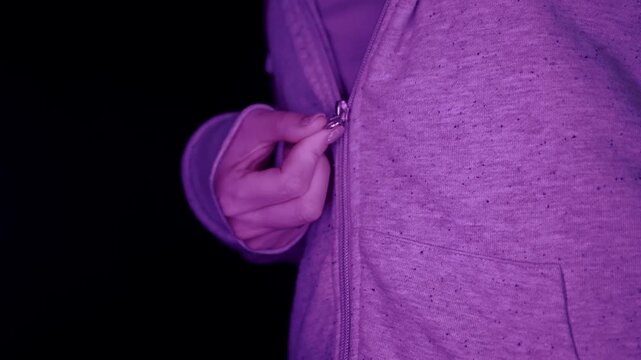 A close-up view captures a woman fastening the zipper of a grey hooded sweatshirt under purple lighting. Side view. Concept of casual fashion, getting dressed, and preparing for cold weather.