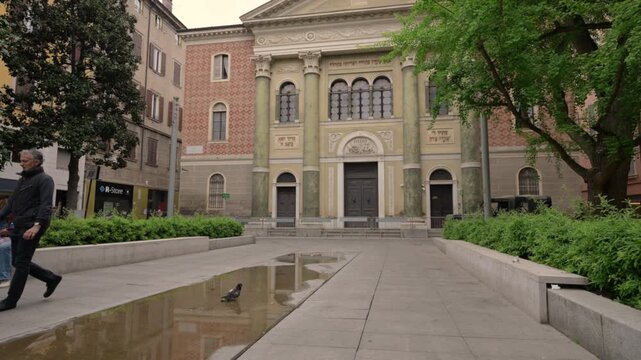 Modena, italy, april 12, 2026. Majestic facade of the synagogue of modena, italy, with people walking in the square. A puddle reflects the building and the sky