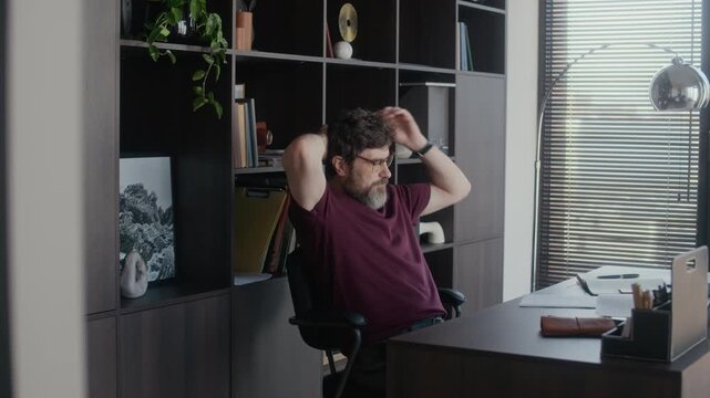 Middle aged male writer working at desk in home office, taking small pause then taking papers with plot plan