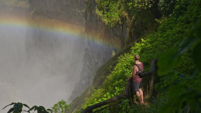 Young male tourist contemplating magnificent Victoria Falls and rainbow from scenic observation point in lush green jungle, appreciating Mosi oa Tunya natural wonder