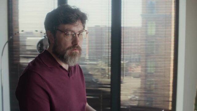 Medium close up of mature Caucasian male editor reading short story draft then looking at camera during workday in modern office