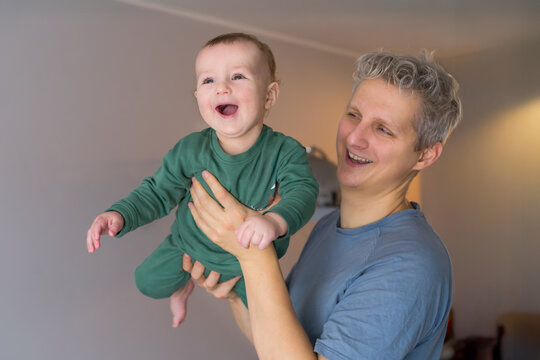 Father and toddler son having fun. Young father lifts his joyful baby in a cozy room. Baby laughing, showing delight. They enjoy their time together at home. Dad playing with baby, happy childhood.