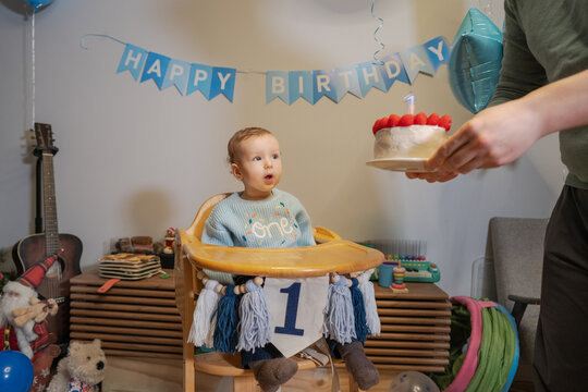 Baby boy sits in high chair looking surprised while father holds a birthday cake with candles. Colorful decorations fill the living room. Family gathers to celebrate first birthday occasion.