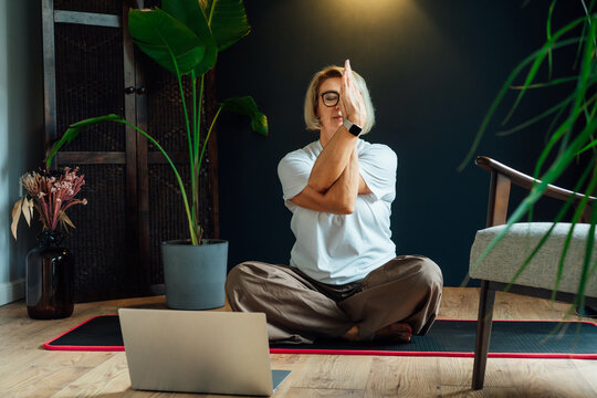 Middle aged woman practicing online yoga workout using laptop in modern Scandi dark interior home. Healthy Lifestyle. Sport activity for mature wellbeing. Fitness online training. Mindful movement