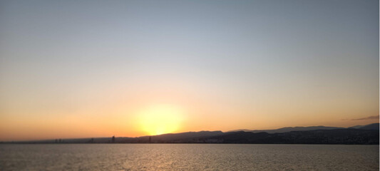 Obraz premium Sunrise over mountains, sea, and city with a glowing orange sky.