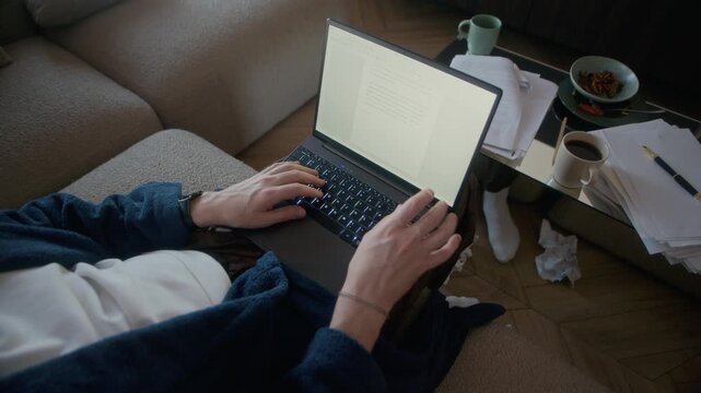 High angle view of unrecognizable male author sitting relaxed on coach in messy living room, writing novel on laptop