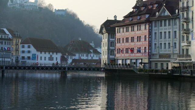 Local people walking across the historic Reussbr&uuml;cke bridge, featuring traditional painted guild houses and Swiss flags along the riverfront in Lucerne.