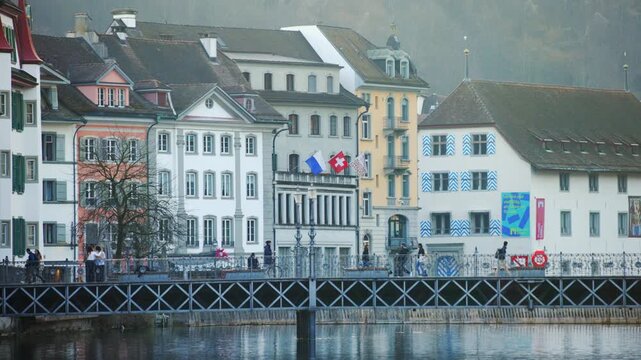 Local people walking across the historic Reussbr&uuml;cke bridge, featuring traditional painted guild houses and Swiss flags along the riverfront in Lucerne.