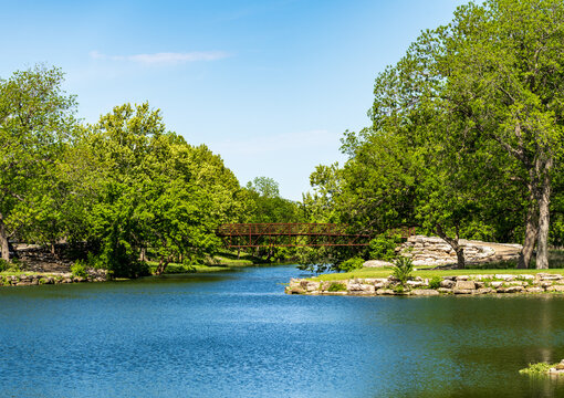 A tranquil view of a clear lake reflecting the surrounding greenery and a small bridge. Serene waterscape in Legacy Hills Park, Georgetown, Texas.