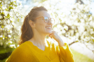 Backlit profile portrait of a woman wearing glasses and a yellow sweater, looking peacefully at blooming white flowers in a sun-drenched spring garden. © Alliance