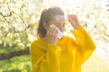 Obraz premium A woman in a yellow sweater and glasses uses a tissue to manage her hay fever symptoms while surrounded by blooming spring orchard trees.