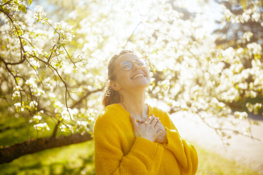 A peaceful woman in a yellow knit sweater closes her eyes and presses her hands to her chest, surrounded by white flowering trees in the golden hour light.