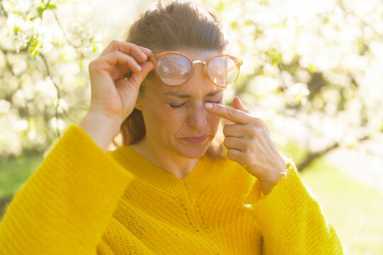 A woman in a yellow sweater rubs her itchy eye while standing among blooming trees, depicting common seasonal allergy challenges and pollen sensitivity.