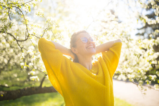 A young woman in a yellow sweater and clear glasses smiles with her hands behind her head, enjoying a beautiful sunny day among white spring blossoms.