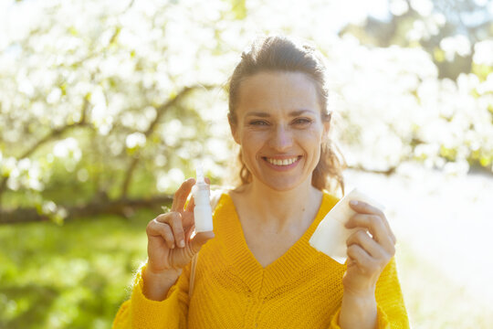 A smiling woman in a yellow sweater holds her nasal spray and tissue, showing effective management of seasonal hay fever while enjoying the spring blossoms.