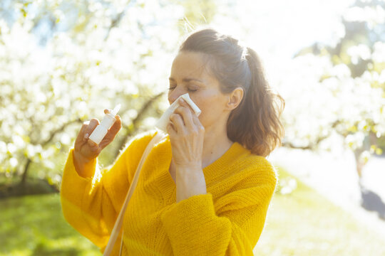 A woman deals with seasonal hay fever symptoms, using a tissue and nasal spray while standing in a beautiful but pollen-filled spring garden.