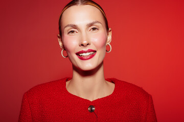 Smiling woman with red lipstick and red jacket poses on red background. Close-up portrait with hoop earrings and natural makeup. Concept of beauty, fashion, and confidence. © SHOTPRIME STUDIO