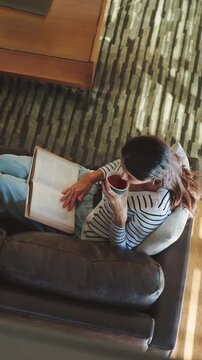 Drink, reading book and above of woman on sofa for literature, hobby and relax on weekend. Home, rest and person on couch with beverage for story, novel and chapter with calm, peaceful and me time
