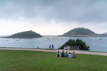 Naklejka premium People enjoying coastal views from a green lawn overlooking Santa Clara Island in San Sebastian, Spain.