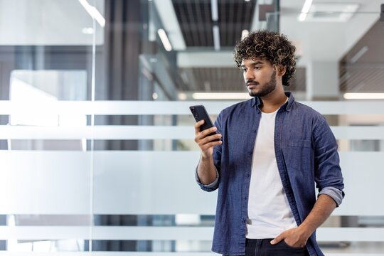 Young Indian man standing in an office building, holding his hand in his pocket and using his phone
