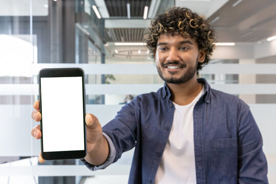 Close-up photo of a smiling young Indian man in an office, showing a white screen mockup of a mobile phone to the camera